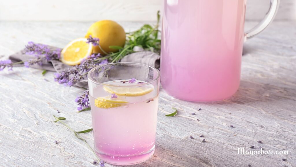a pitcher of pink lemonade next to a glass with lemons and lavender